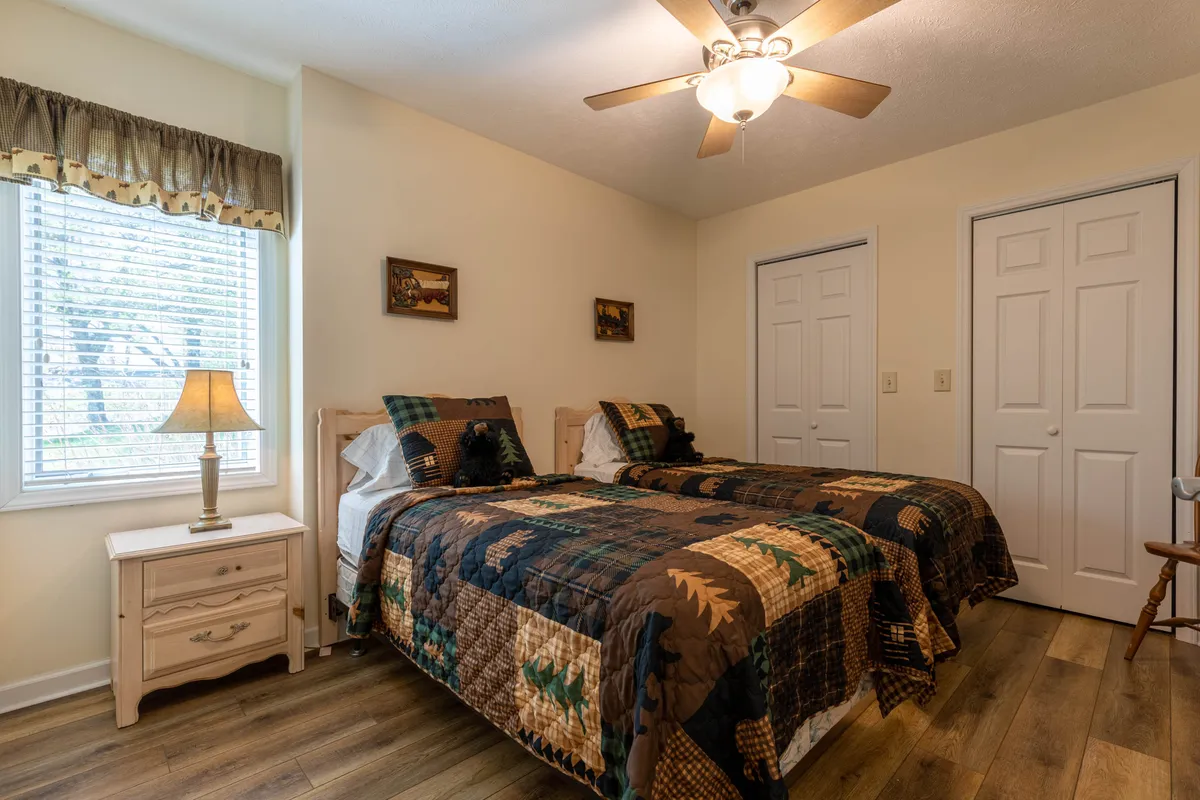 A cheerful bedroom with twin beds, a window with blinds, and a nightstand with a lamp. The space feels inviting and practical—ideal for children or friends, and naturally lit for a restful ambiance.