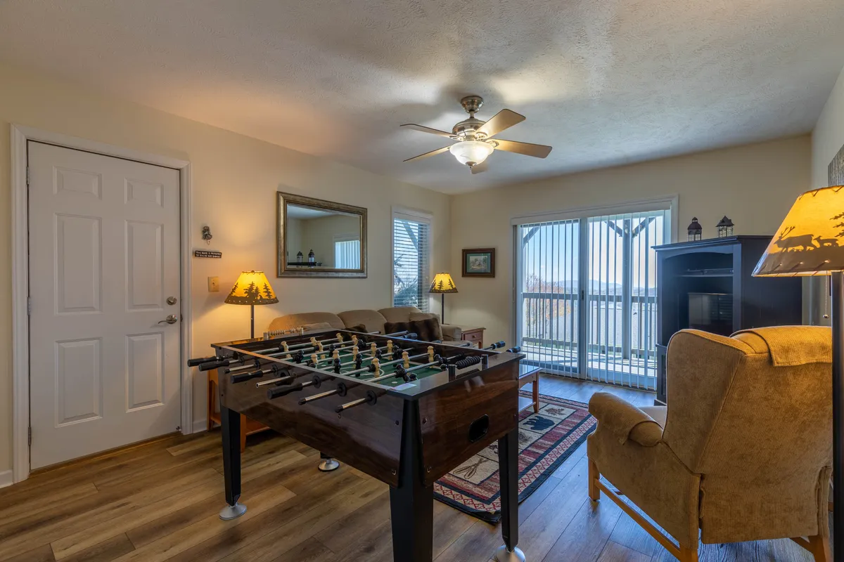 This inviting interior scene centers on a foosball table flanked by comfortable seating and large windows streaming with natural light. The space is clearly designed for both leisure and lively activity, with the foosball table front and center for friendly competition. A decorative lamp and warm furnishings add to the cozy, fun atmosphere.