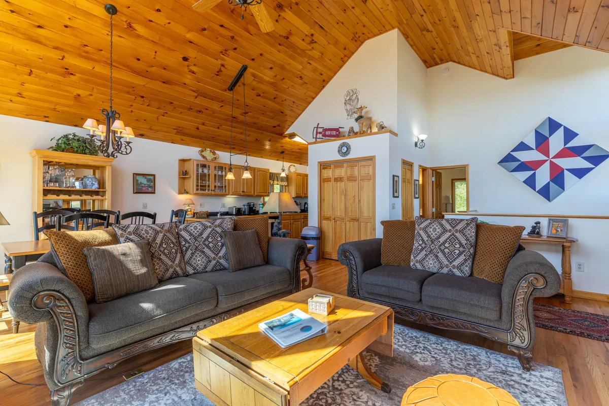 This cozy living room features inviting sofas arranged for socializing, a rustic wooden ceiling, a central coffee table, and mountain-themed wall art. Large windows in the background hint at the natural scenery just beyond, creating a seamless indoor-outdoor connection ideal for relaxing with your pets after a day of exploration.