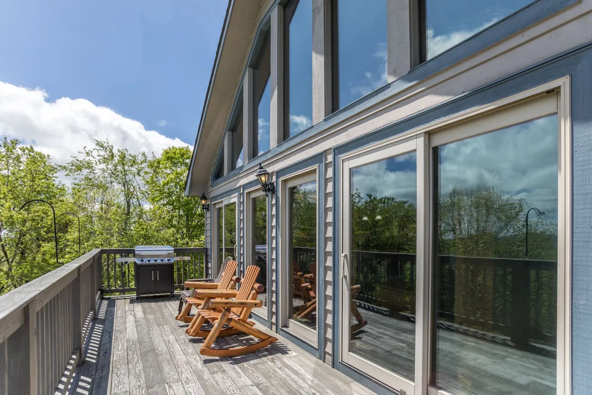 This image displays a spacious wooden deck featuring multiple Adirondack chairs, an outdoor grill, and large windows that connect the deck to the house. The deck is surrounded by a tranquil natural setting, perfect for enjoying the outdoors.