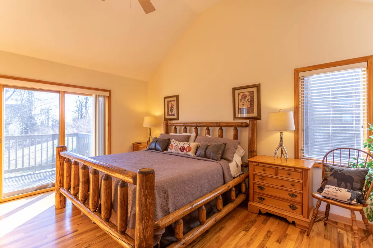 A warmly lit bedroom featuring a log bed placed near large windows, a nightstand, and rustic artwork on the wall. Natural light pours in, creating a peaceful atmosphere ideal for relaxation or enjoying a quiet morning coffee in bed.