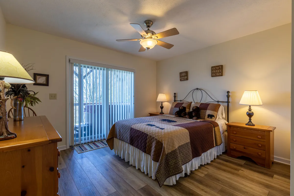 A cozy bedroom with a decorative quilted bed, flanked by nightstands with lamps, and a sliding glass door that leads out to a private balcony. The room is bathed in natural light, embodying comfort and privacy.