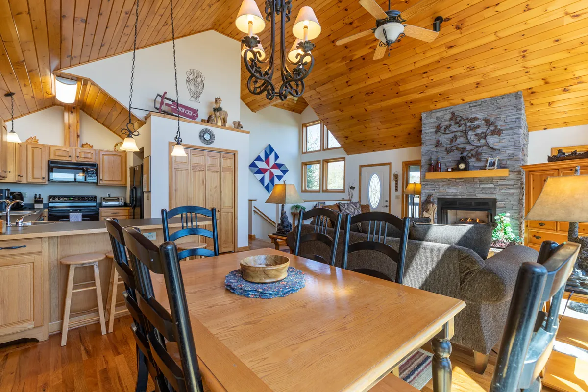 This image features a large wooden dining table set beside a striking stone fireplace and beneath a wood-paneled cathedral ceiling. A chandelier adds a touch of elegance, while the overall ambiance is cozy and inviting—perfect for family dinners, celebrations, and relaxing evenings by the fire.