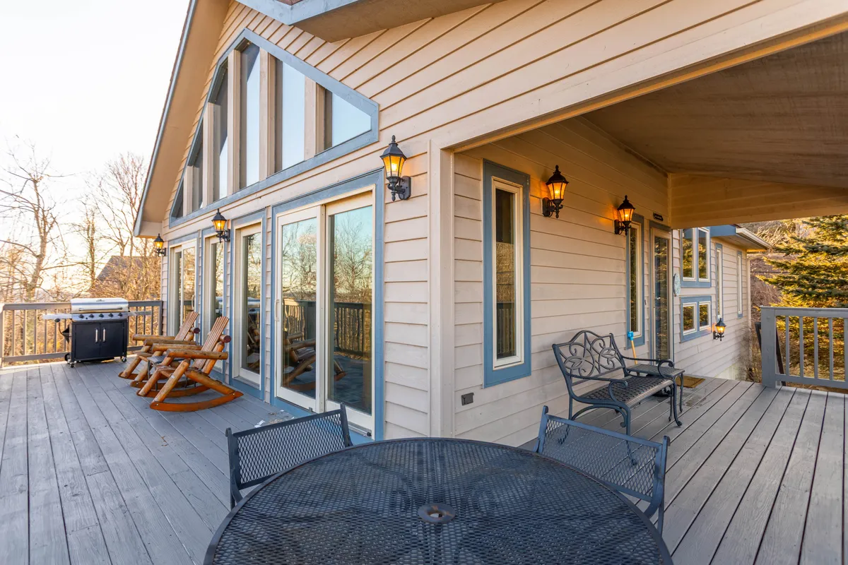 The photo shows a cozy deck space with large glass windows, rocking chairs, an outdoor dining table set up for meals, and charming lantern-style lights. The setting is ideal for alfresco dining and casual gatherings, with a warm, inviting atmosphere.