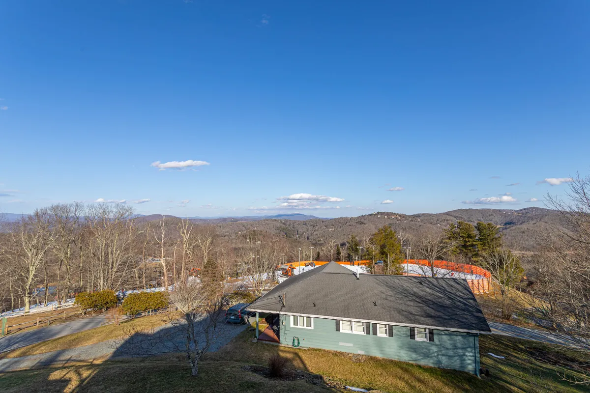 This exterior shot features the green house nestled against a dramatic mountain range, under a clear blue sky. The wide perspective highlights both the home and the striking natural landscape, emphasizing the property's prime location for outdoor adventure.