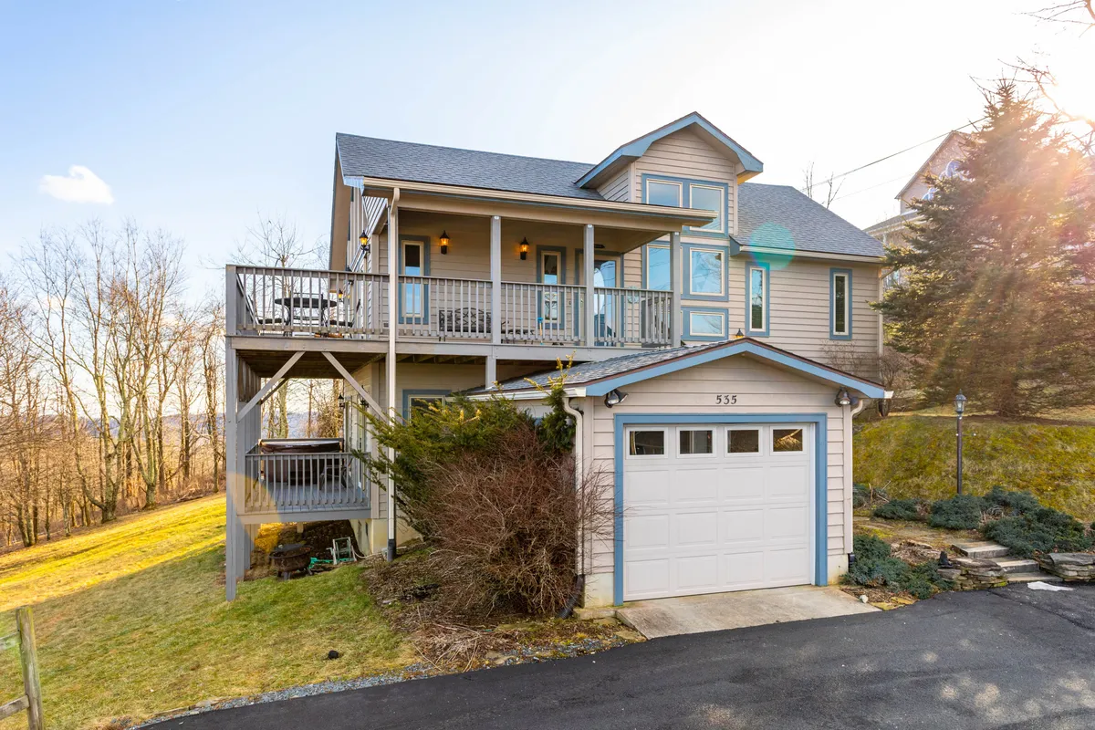 The image shows the welcoming exterior of a pristine single-family mountain home perched on a gentle slope, featuring a spacious balcony perfect for soaking in panoramic sunset views. The front door and garage are visible, with the balcony set for morning coffee or evening relaxation overlooking the Blue Ridge Mountains, creating an inviting scene for outdoor gatherings or quiet moments amid nature.